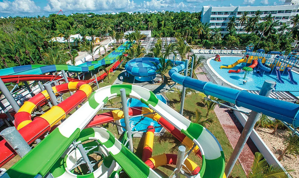 Aerial view of a vibrant water park at a tropical resort, featuring colorful winding water slides, swimming pools with children's features, palm trees, and a hotel building in the background under a sunny sky.