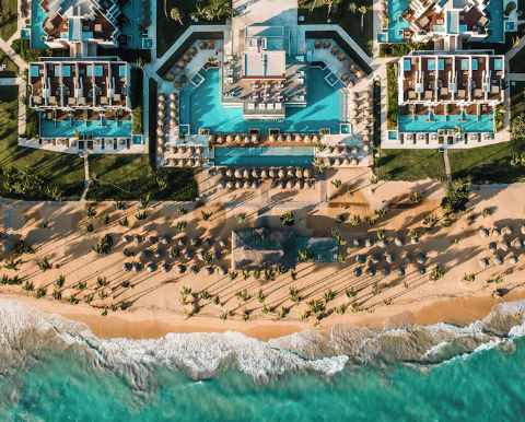 Top-down aerial view of a symmetrical luxury beach resort in Punta Cana, showing pools, private villa sections, and a sandy beach with palapas and palm trees next to the turquoise sea.
