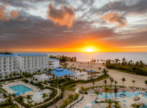 Aerial view of a tropical beachfront resort at sunrise featuring white buildings, swimming pools, palm trees, and a sandy beach by the ocean