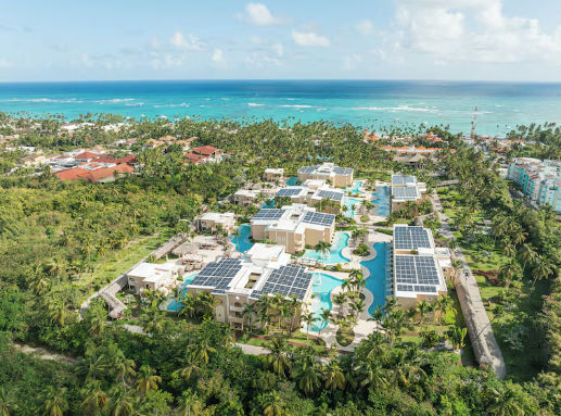 Aerial view of a tropical beach resort with solar panels on roofs, swimming pools, and the ocean in the background