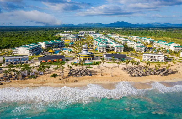 Aerial view of a large tropical beach resort complex. In the foreground is the turquoise ocean with white waves and a wide sandy beach dotted with clusters of thatch-roofed umbrellas and palm trees. Behind the beach is the extensive resort, featuring multiple multi-story white buildings with green roofs, swimming pools, a central lighthouse-style tower, and surrounding lush green landscaping. A vast green forest leads to a range of blue mountains in the background under a blue sky with puffy white clouds.