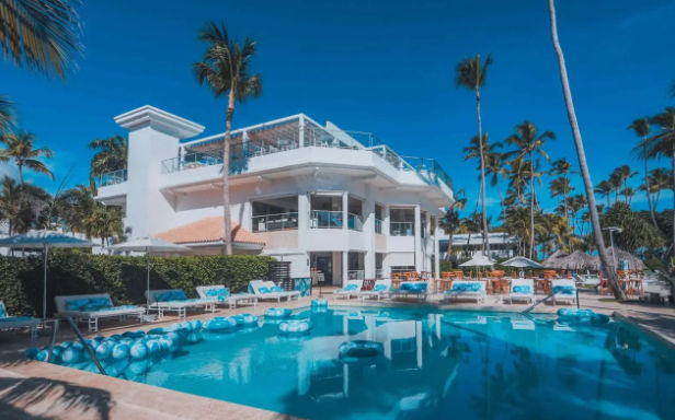 A modern white luxury beachfront resort hotel in Punta Cana with a large swimming pool and palm trees under a blue sky.