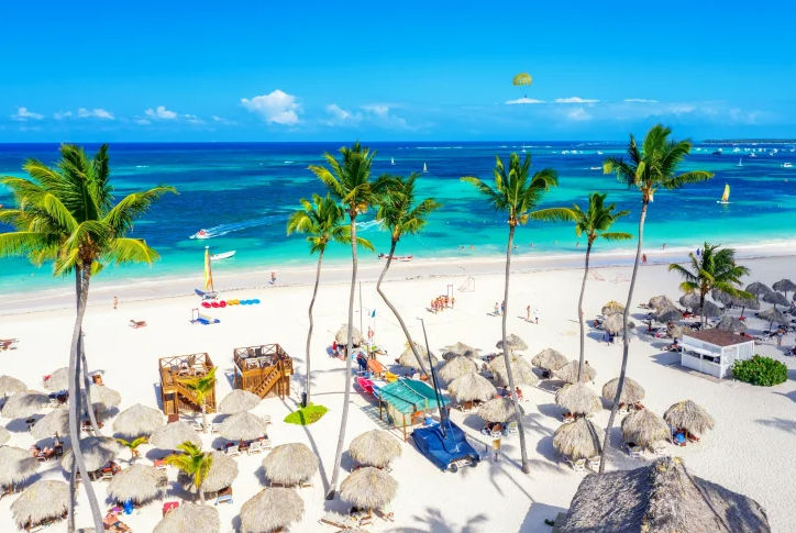 Aerial view of a beautiful tropical beach with white sand, tall palm trees, thatched umbrellas, and bright turquoise ocean water