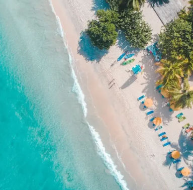 Drone view of a Punta Cana beach with turquoise water, white sand, lush trees, striped umbrellas, lounge chairs, and people by a lifeguard stand