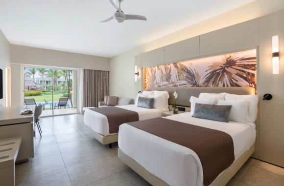 Modern resort room with two beds, tropical wall art, and a sliding glass door opening to an outdoor patio with pool access.