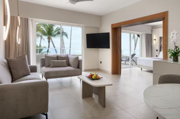 Interior view of a modern hotel suite with a living area, bedroom, and balcony overlooking palm trees and the ocean.