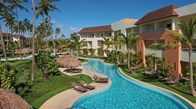 Elevated view of a luxurious tropical resort with a winding lazy river pool, palm trees, and sunbeds.