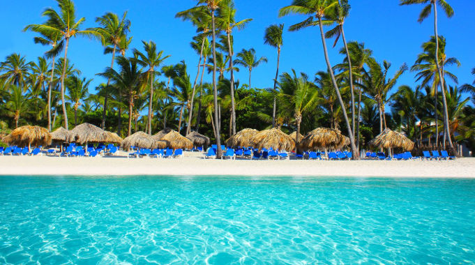 Tropical beach with white sand, palm trees, thatched umbrellas, blue lounge chairs, and clear turquoise water