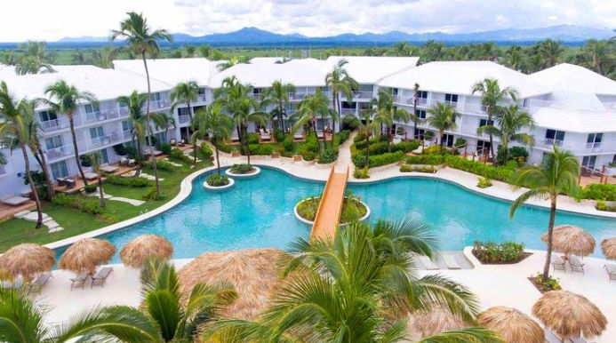 An elevated, wide view of a luxury tropical resort with white buildings and extensive swimming pools, connected by a footbridge, surrounded by palm trees and palapas, with mountains in the background under a cloudy sky.