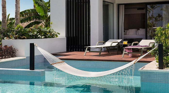 Luxurious modern swim-up resort suite with a large rope hammock suspended over the clear blue pool water, leading to a private deck with two chaise lounges and open bedroom doors, all surrounded by lush tropical landscaping.
