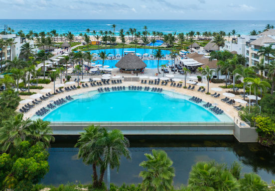 Aerial view of a luxury tropical resort with large swimming pools, lounge chairs, palm trees, and ocean in the background