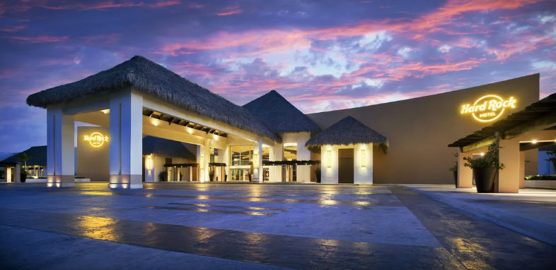 Illuminated entrance of a Hard Rock Hotel at dusk with a large thatched-roof porte-cochère and dramatic sky.