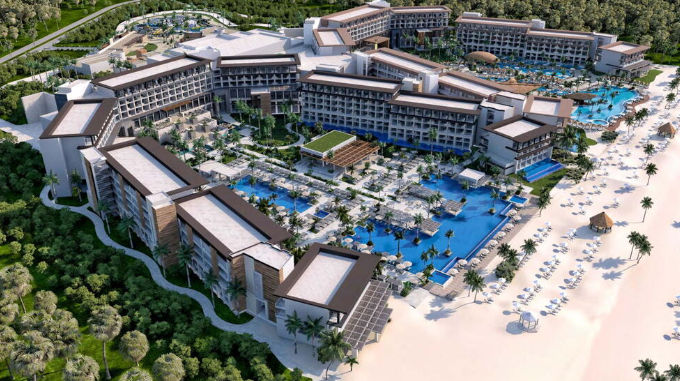 Aerial view of a sprawling modern luxury beachfront hotel complex with massive pools, palm-lined courtyards, and a sandy beach with sun loungers.