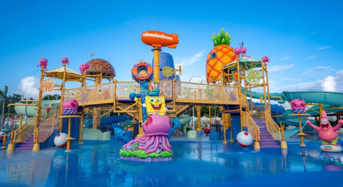 Vibrant outdoor Nickelodeon-themed water park, Aqua Nick, featuring SpongeBob SquarePants on a jellyfish and Patrick Star under a clear blue sky.