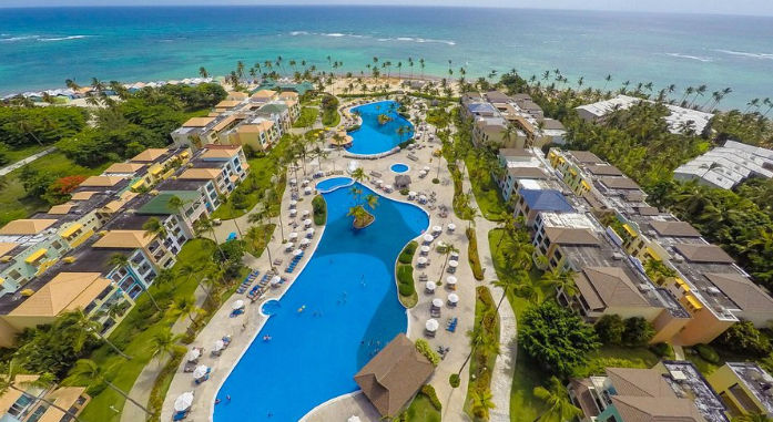 Aerial view of a tropical beach resort with massive curved swimming pools, palm trees, and buildings near a sandy beach and clear turquoise ocean water.