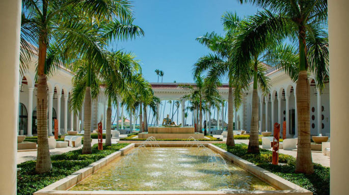 Luxurious resort courtyard with a central water fountain, lined with tall palm trees and elegant white arched colonnades under a clear blue sky.