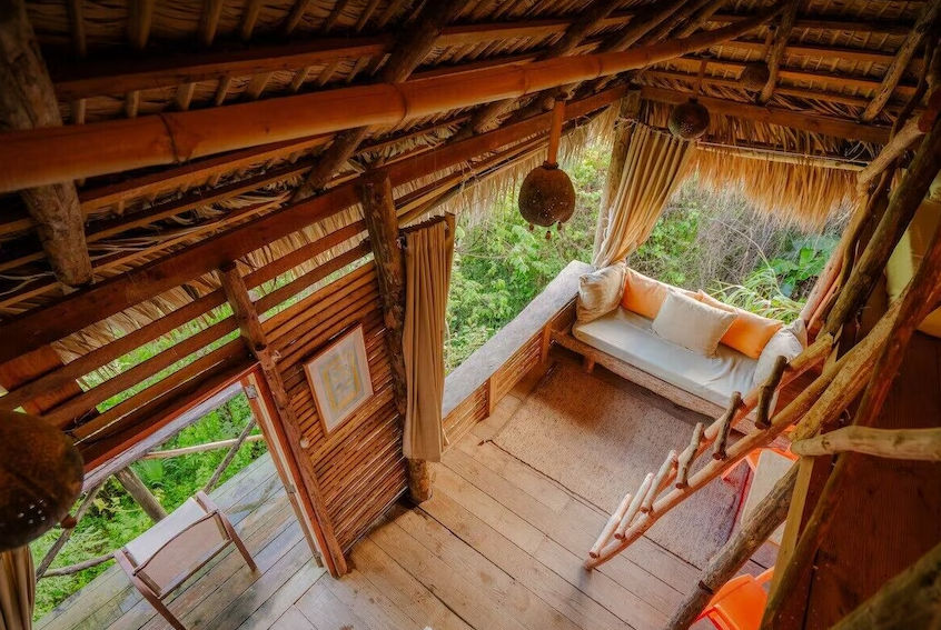 Rustic eco-lodge cabin with a thatched roof, a small plunge pool, and a wooden lounge chair on a stone patio, overlooking a lush green valley and the ocean.