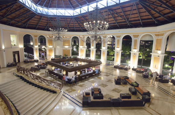 Aerial view of a luxurious hotel lobby atrium with a central square bar, an intricate wooden domed ceiling with skylights, and a grand curved staircase, showing patrons and seating areas.