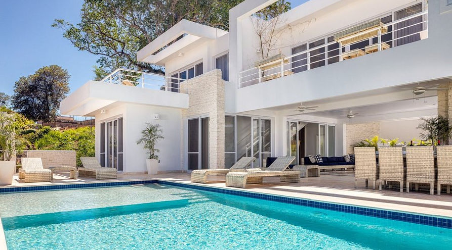 Exterior view of a modern white luxury villa featuring a large private swimming pool, wicker lounge chairs, and a covered outdoor patio under a clear blue sky.