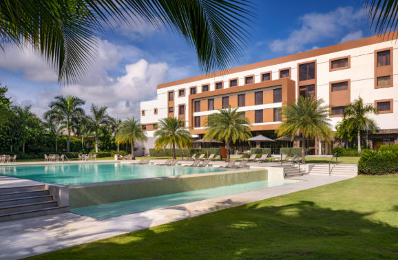 Modern luxury hotel resort with a large swimming pool, palm trees, and green lawns under a blue, partly cloudy sky.