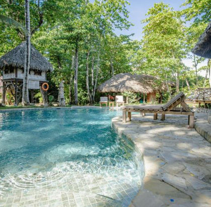 Outdoor swimming pool surrounded by lush tropical trees, featuring rustic thatched-roof cabanas, a stone deck, and a wooden lounge chair by the water.
