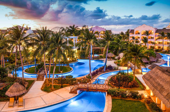 Aerial twilight view of a luxury tropical resort with a winding lazy river pool, illuminated paths, wooden bridges, palm trees, and multi-story buildings under a dramatic purple and blue sky.