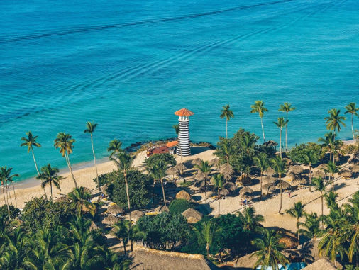 High-angle drone view of a tropical Caribbean beach with turquoise water, a striped lighthouse, and numerous palm trees over thatched huts.
