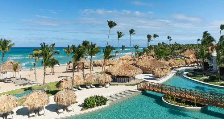 Luxury beachfront resort with a swimming pool, wooden bridge, thatched umbrellas, and palm trees near the ocean