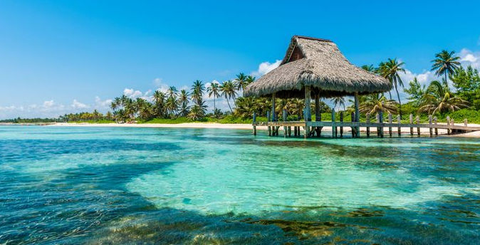 Tropical beach with a wooden pier and thatched-roof hut over clear blue water under a clear sky