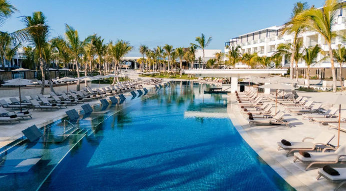 A long infinity swimming pool at a luxury tropical resort flanked by palm trees, rows of lounge chairs, and a modern hotel building.