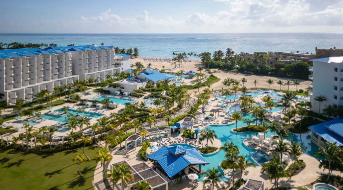 Aerial view of a sprawling beachfront luxury resort with multiple connected swimming pools, white hotel buildings with blue roofs, palm trees, and the ocean under a cloudy sky.