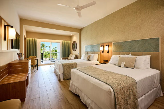 Bright hotel room with two beds, wooden furniture, ceiling fan, and a balcony with a tropical palm tree view