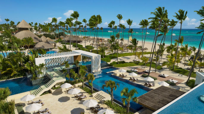 Aerial view of a luxury tropical resort with multi-tiered swimming pools, white umbrellas, palm trees, and a sandy beach facing the turquoise ocean.