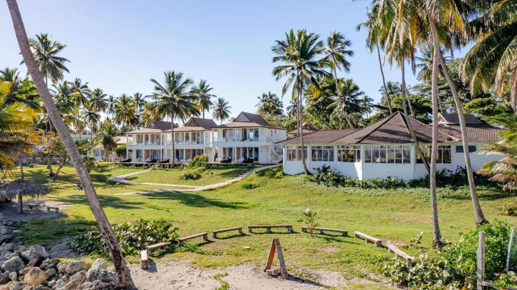Una vista bañada por el sol del complejo turístico de Las Canas Beach Retreat, compuesto por edificios blancos situados entre una densa arboleda de imponentes palmeras, que cuenta con cuidados jardines verdes, un camino de piedra serpenteante, una costa r