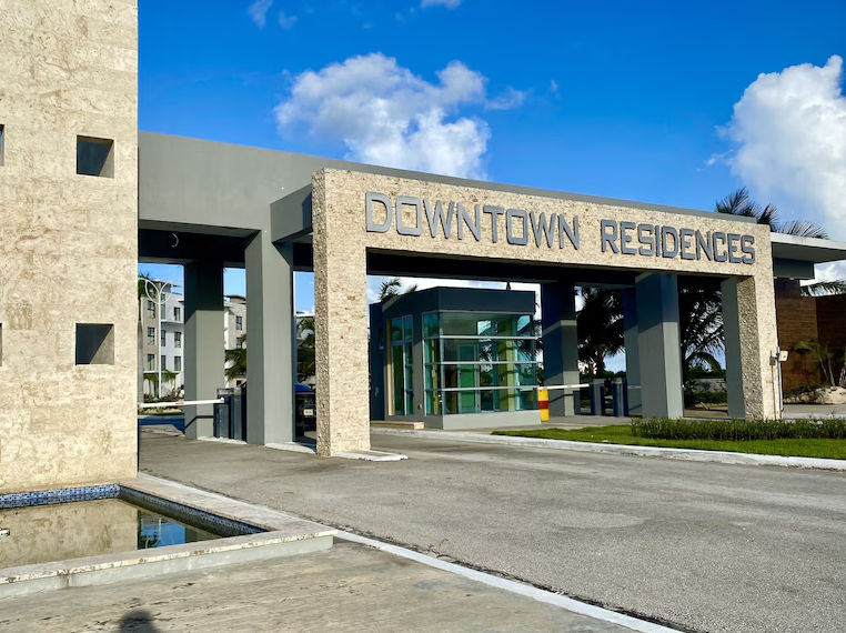 Entrada principal moderna al complejo residencial DOWNTOWN RESIDENCES con columnas de piedra, garita de seguridad y barreras de acceso vehicular bajo un cielo azul soleado.