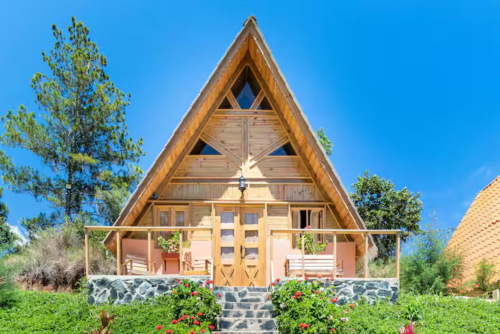 A charming, rustic two-story mountain cottage with bright orange stucco and rough stone facade, surrounded by pine trees. It features a large timber-framed front porch and a red A-frame roof.