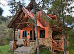 A charming, rustic two-story mountain cottage with bright orange stucco and rough stone facade, surrounded by pine trees. It features a large timber-framed front porch and a red A-frame roof.