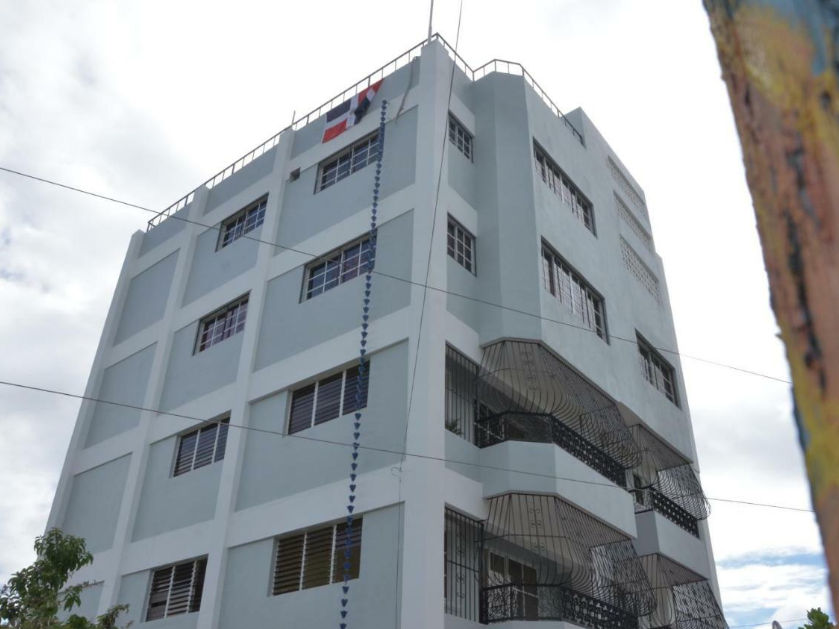 Low-angle shot looking up at the facade of the Hotel Quinto Nivel RD, a six-story building painted light grey. The image shows windows with security bars, balconies on the fourth and fifth floors with decorative black wrought-iron railings, and a Dominica