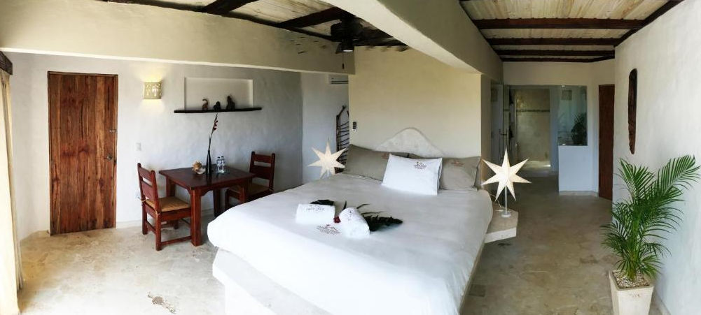 Vista panorámica de la fachada de Cabrera Chalet en República Dominicana, mostrando su diseño arquitectónico de estilo boutique con paredes de piedra natural, balcones, palmeras y un cenador con techo de paja bajo un cielo azul despejado.