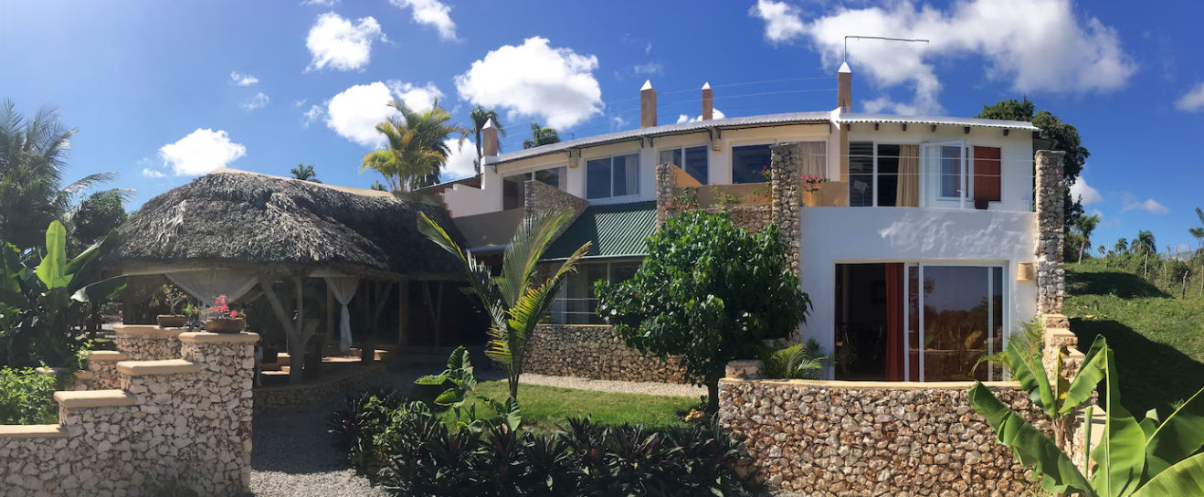 Vista panorámica de la fachada de Cabrera Chalet en República Dominicana, mostrando su diseño arquitectónico de estilo boutique con paredes de piedra natural, balcones, palmeras y un cenador con techo de paja bajo un cielo azul despejado.