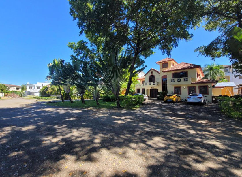 Exterior view of B&B Pavillion villa in Sosua, Dominican Republic, featuring tropical traveler's palms, large shade trees, and a parking area under a clear blue sky.