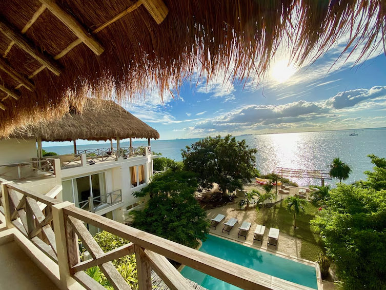 Vista desde un balcón con techo de paja que muestra un complejo hotelero tropical frente al mar, con una piscina, camastros, vegetación abundante y un océano tranquilo bajo un cielo soleado.