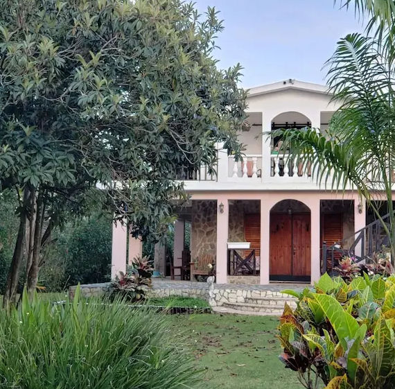 A two-story country house with stone details and wooden doors, surrounded by a lush green garden with trees and tropical plants.