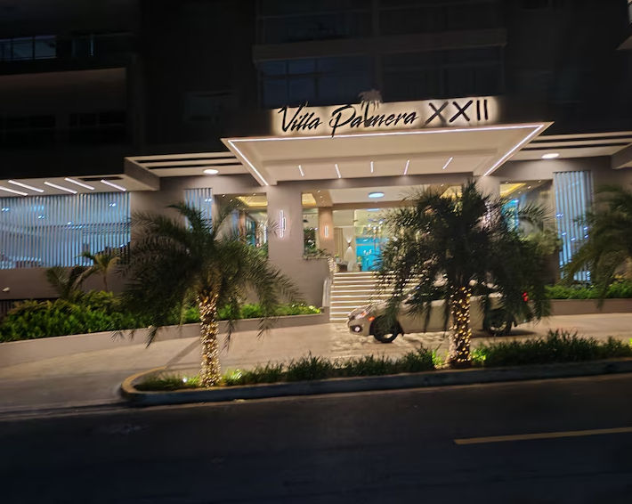 Entrada de la fachada frontal del edificio residencial Villa Palmera XXII por la noche con palmeras iluminadas y un vehículo en la entrada.