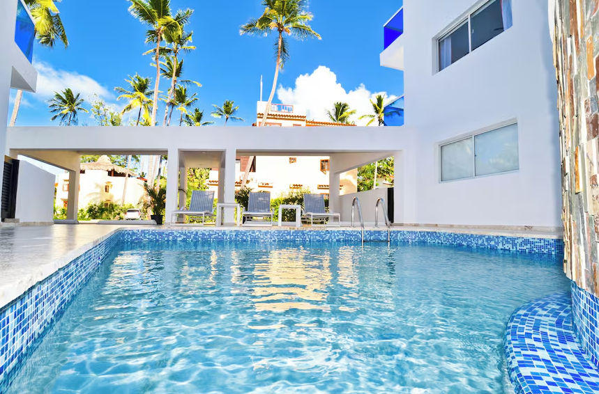 The Sanctuary at Los Corales: Exterior view of a modern white building with bright blue glass balconies, surrounded by tall palm trees under a clear blue sky.