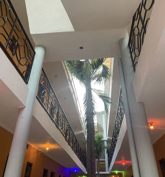 An outdoor corridor of a two-story hotel with yellow walls, brown wooden doors, and a tiled walkway overlooking a small courtyard with palm trees.