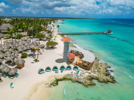 Aerial view of a tropical beach resort featuring a black and white striped lighthouse, thatched umbrellas, and clear turquoise water