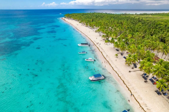 Aerial view of a pristine Caribbean beach in the Dominican Republic with crystal-clear turquoise water, palm trees, and boats anchored off the shore