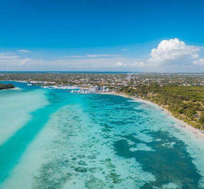Aerial view of a vibrant turquoise bay with shallow waters, a tropical coastline, and a town with a marina in the distance
