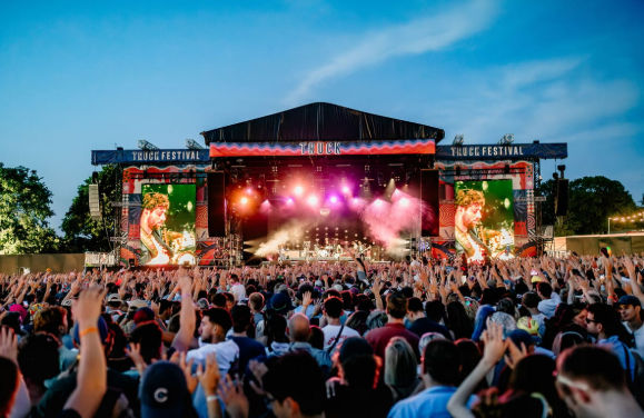 Huge crowd of people with hands in the air in front of an illuminated main stage at the outdoor Truck Festival concert at twilight.
